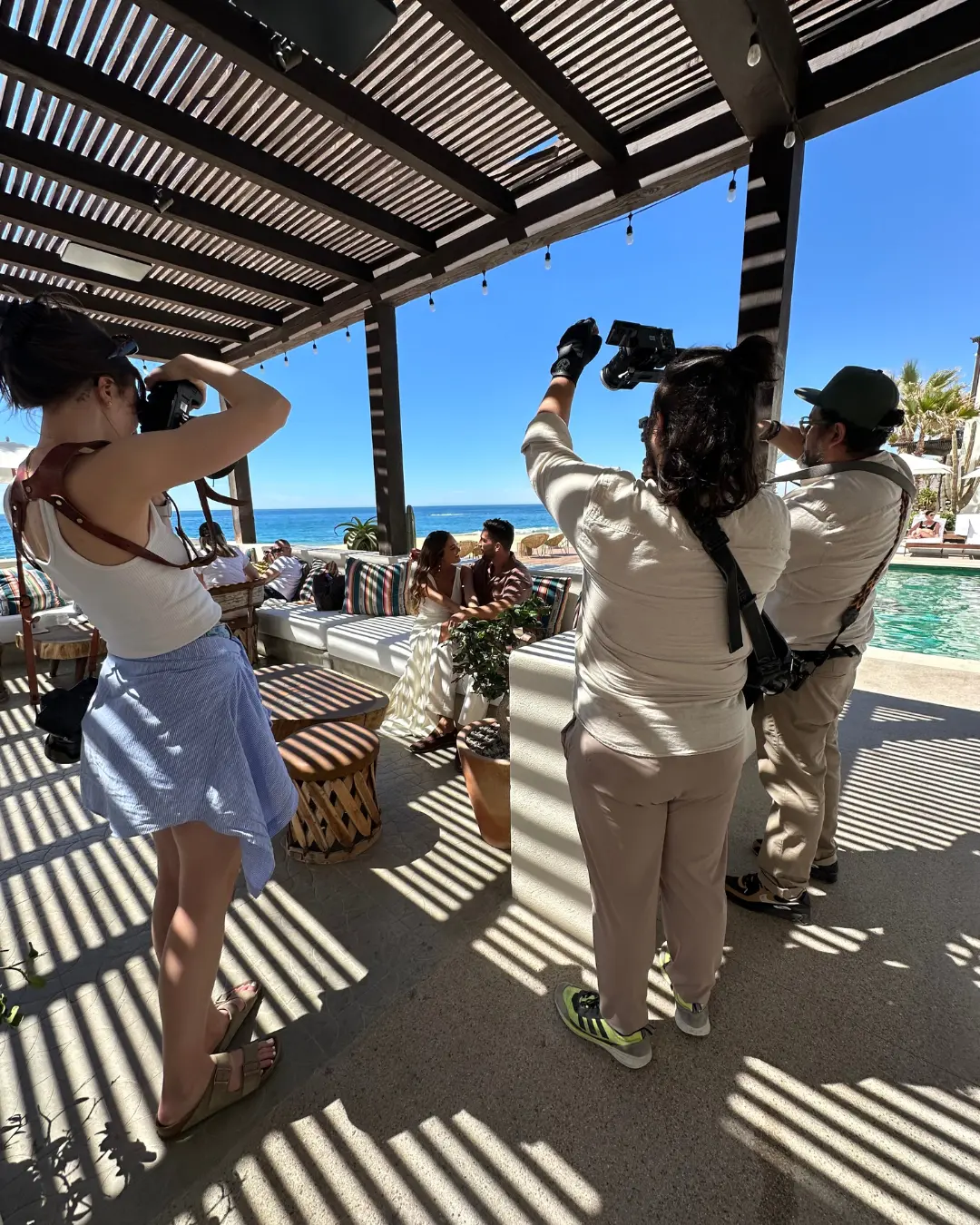 Photographers take pictures of a couple sitting under a shaded pergola next to a pool, with a stunning ocean view in the background. Shadows from the pergola create striped patterns on the ground. Two photographers are closely focusing on the couple, while one adjusts settings.