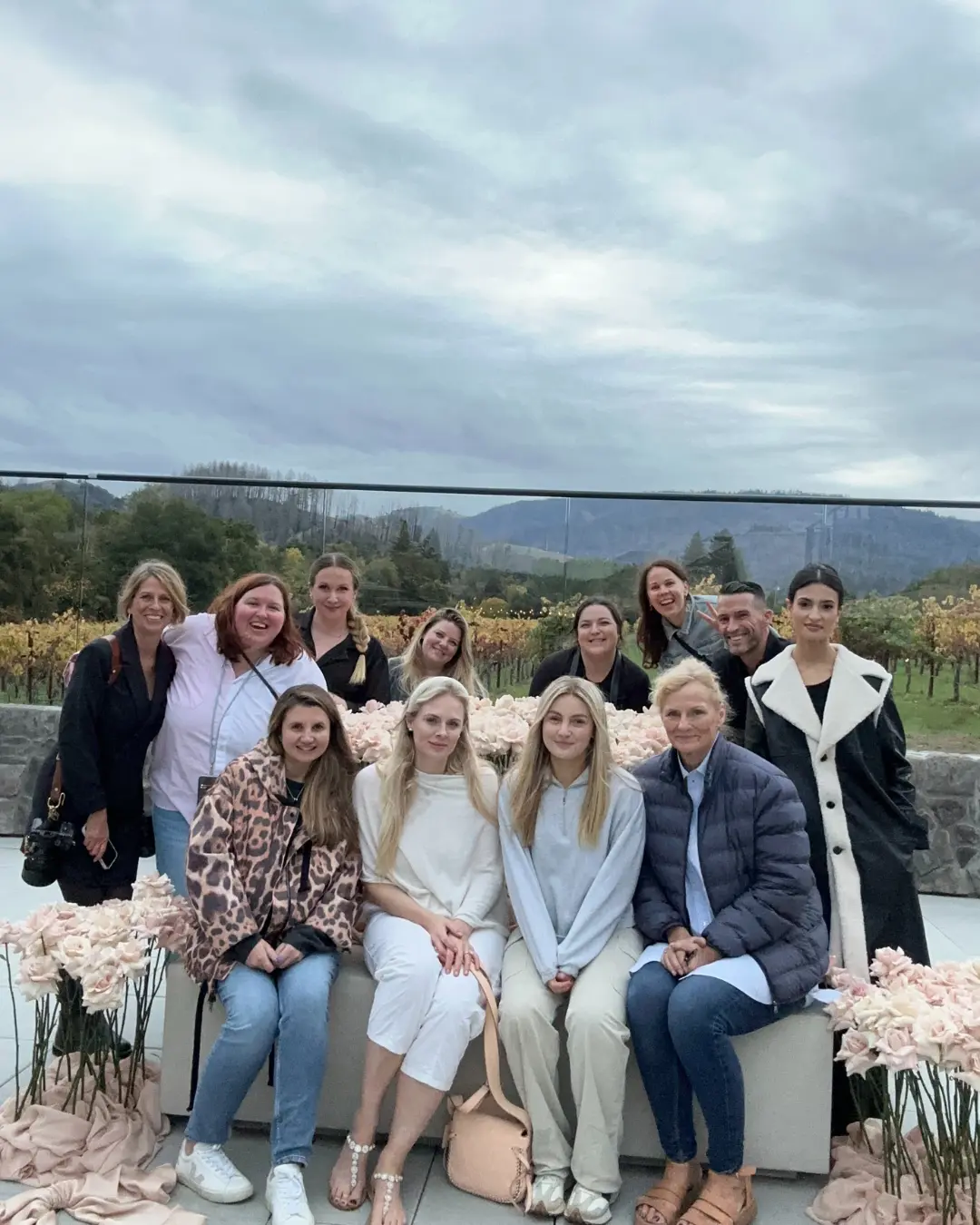 A group of 12 people, consisting of 10 women and 2 men, are gathered outdoors in front of a flower arrangement with a scenic backdrop of vineyards and mountains on an overcast day. The group is smiling and posing for the photo, dressed in casual and semi-formal attire.