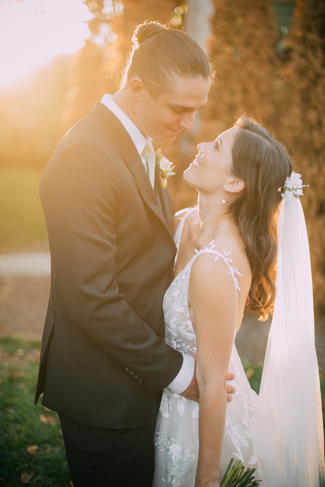 A couple stands outdoors during sunset, dressed in wedding attire. The groom is in a dark suit and has his hair tied back, while the bride is wearing an elegant white gown with floral details and a long veil. They are gazing into each other's eyes and smiling.