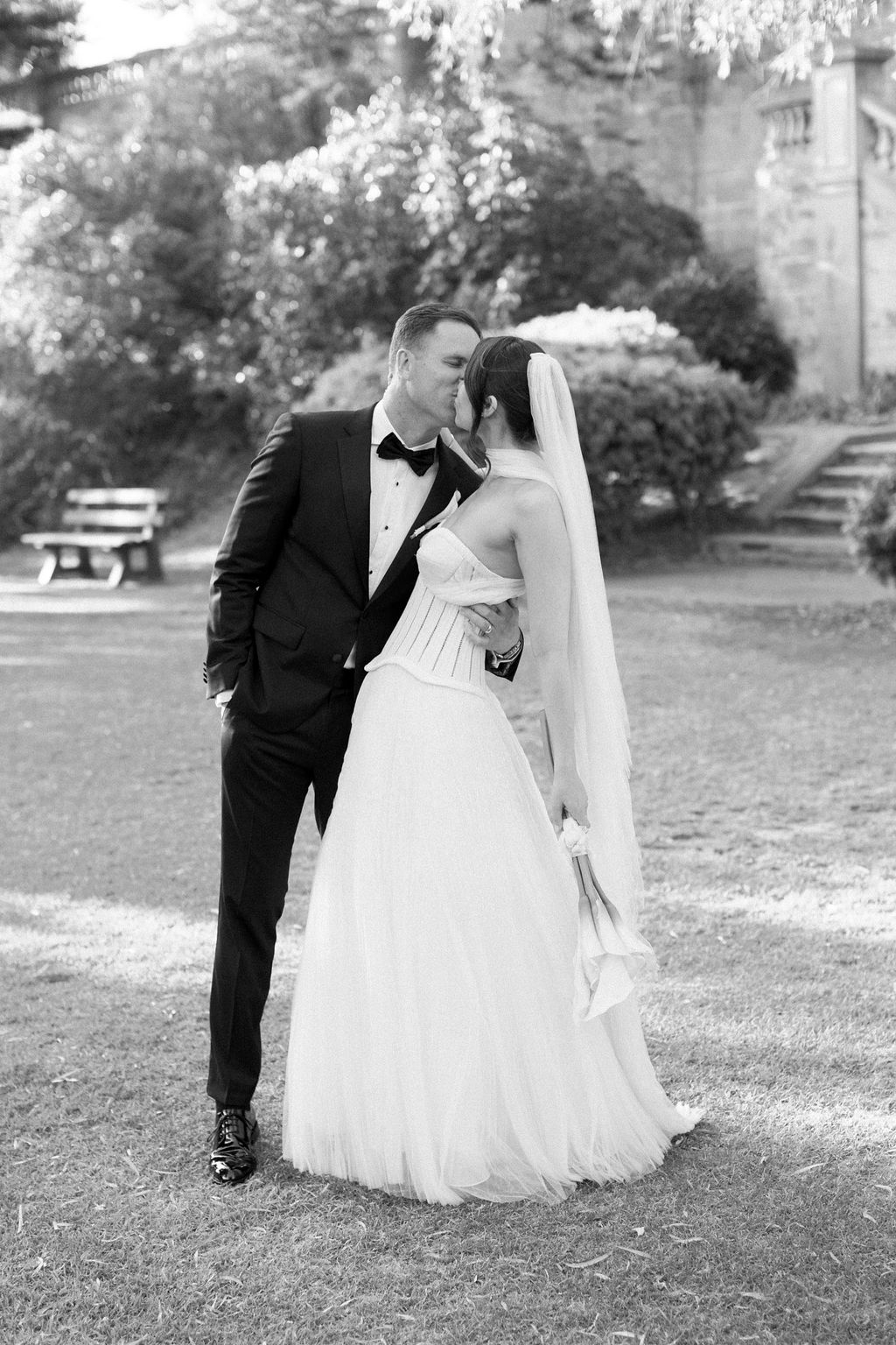 A newlywed couple shares a romantic kiss outdoors. The groom is dressed in a black tuxedo and bow tie, while the bride wears a strapless white wedding gown with a veil. They are surrounded by lush greenery, with a stone bench and stairs in the background.