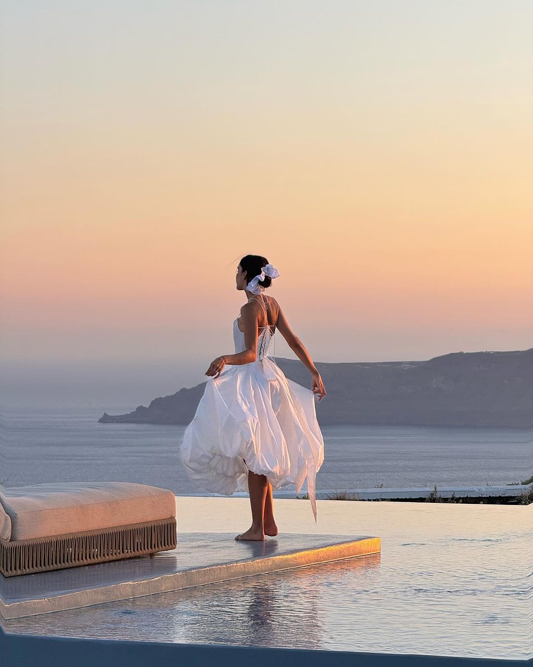 A person wearing a flowing white dress and a white hair bow stands gracefully on the edge of an infinity pool overlooking a large body of water and distant cliffs at sunset. The sky is painted with soft warm hues, reflecting a serene and picturesque scene.