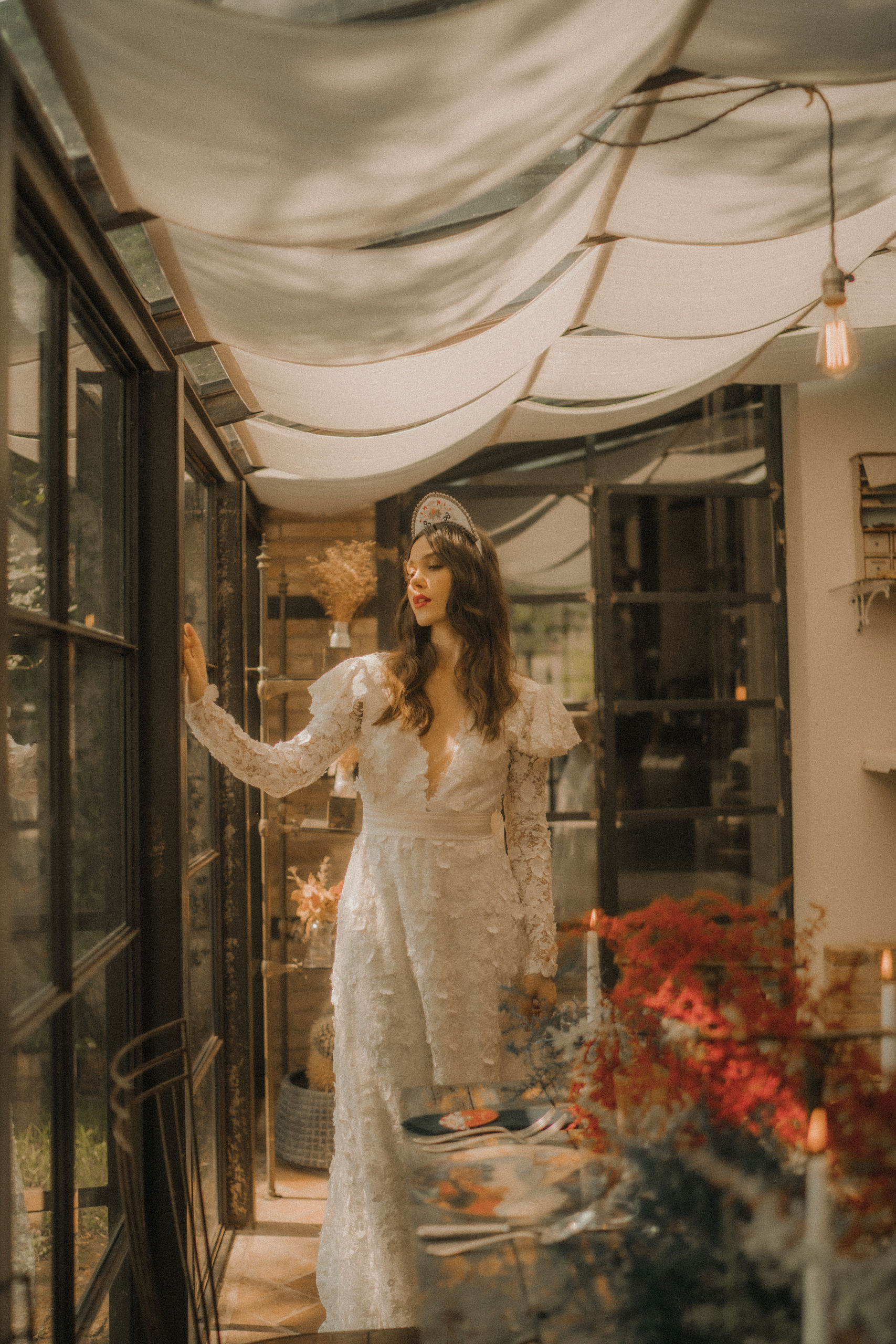 A woman in a long-sleeve white floral dress and tiara stands by a window under a cream-colored canopy. She is looking out, with one hand touching the glass. The setting is warm and decorated with plants and hanging lights.