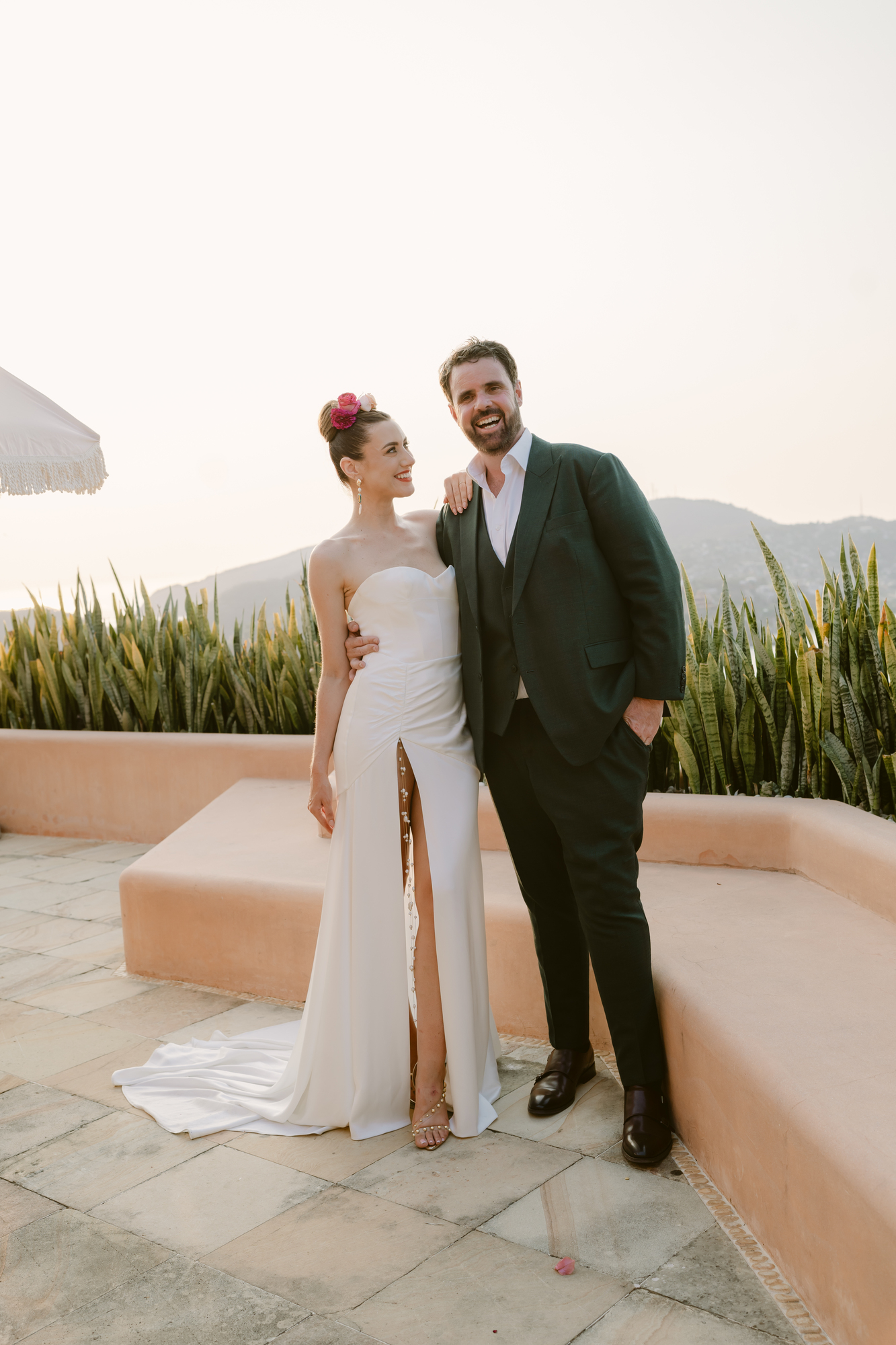 A smiling couple stands together outdoors in formal attire. The woman wears a strapless white dress with a high slit and pink flower hair accessories, while the man wears a dark suit with a vest. They are on a patio with a railing, plants, and hills in the background.