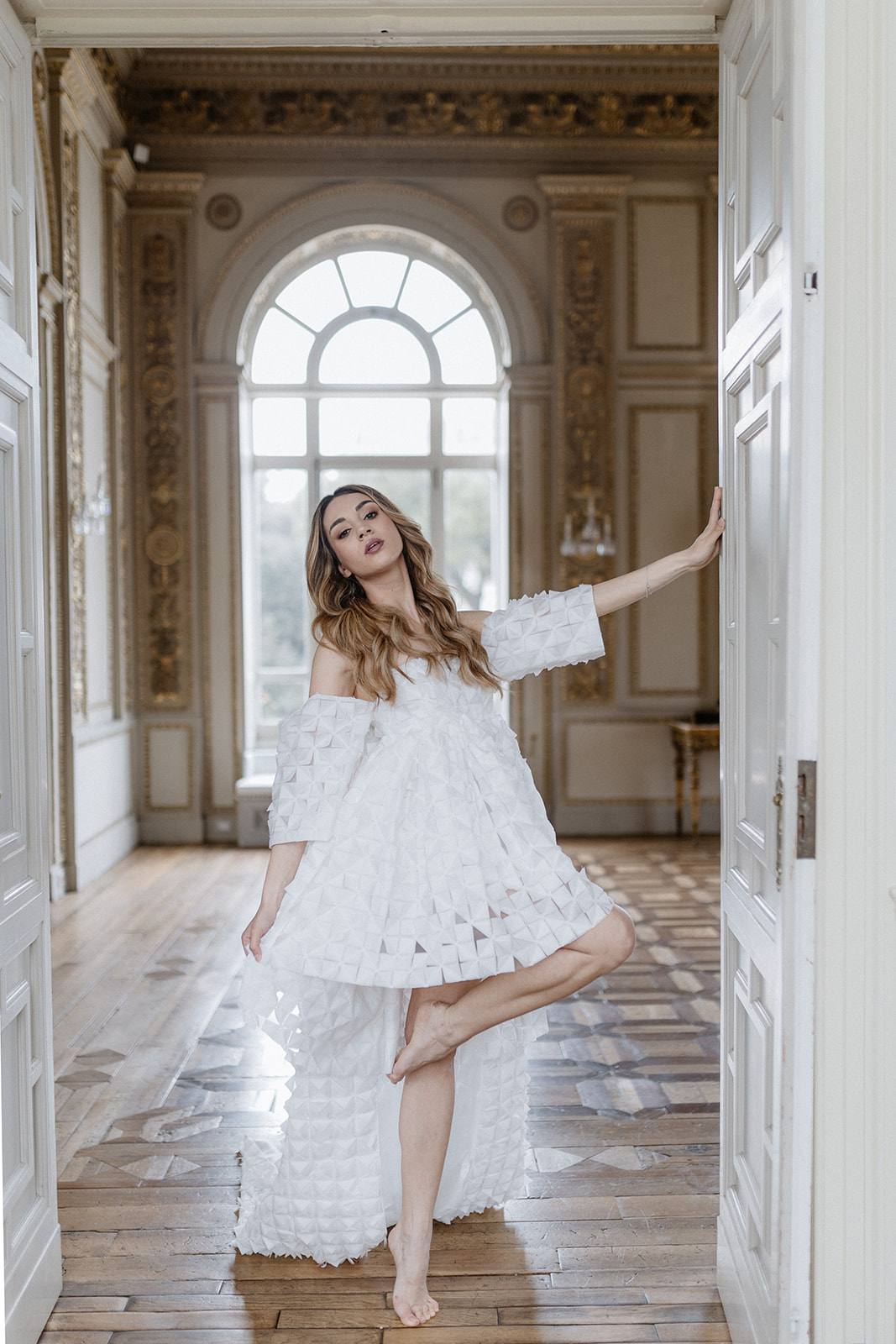 A woman in a textured white off-shoulder dress stands on one foot in a doorway of an ornate room with wooden floors and large windows. She has long hair and is gracefully touching the doorframe with one hand while raising her other hand.