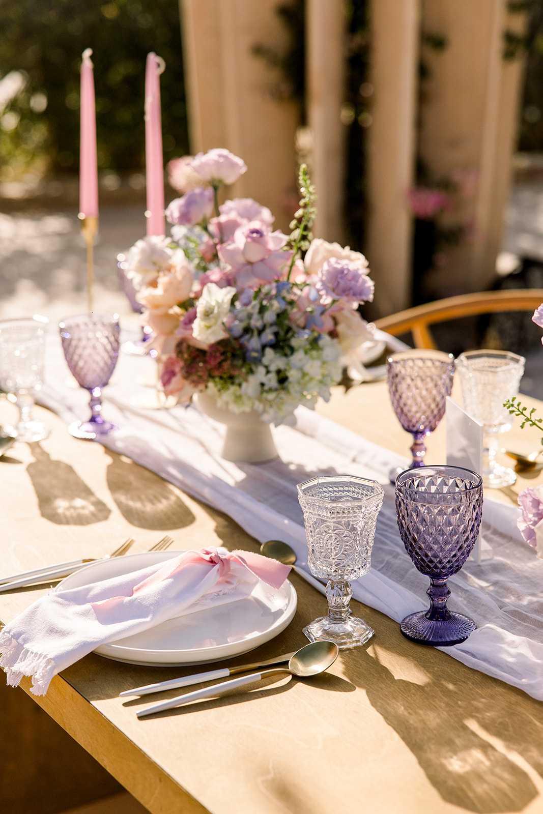 A beautifully set outdoor table with elegant purple and clear glassware, white plates with pink flower napkins, and a centerpiece of white and pastel flowers. Two tall pink candles are in the background, adding a romantic touch to the scene.