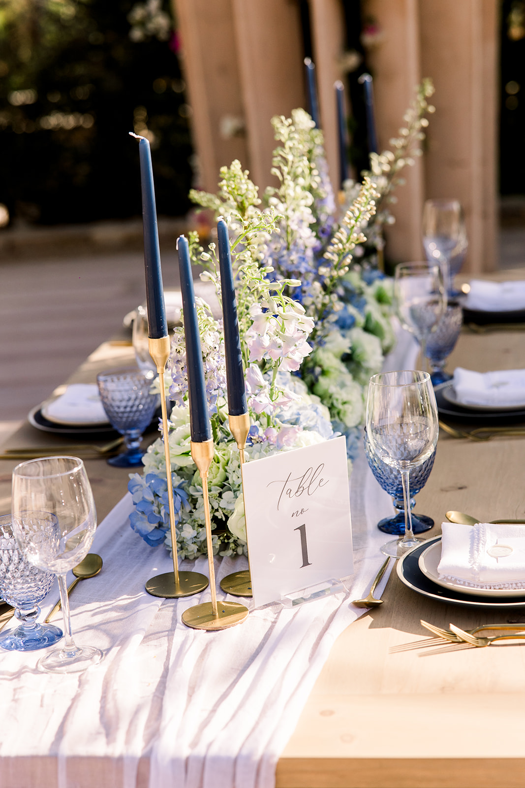 Elegant wedding table setup with a wooden table adorned with a white table runner, tall blue candles in gold holders, blue and white floral centerpiece, crystal glassware, and a "Table No. 1" sign. Plates with elegant napkin settings are arranged neatly.