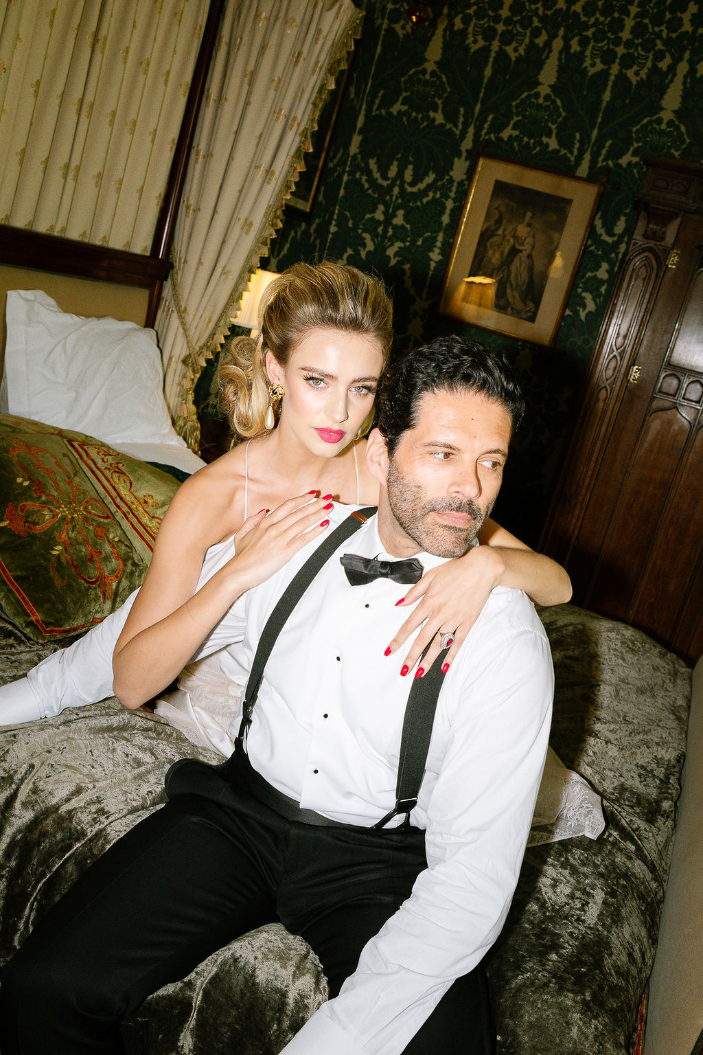 A woman with blonde hair in a white dress sits on a bed, embracing a man in a white dress shirt, black bow tie, and black suspenders. The man has short dark hair and wears a serious expression. They are in a room with ornate wallpaper and antique furniture.