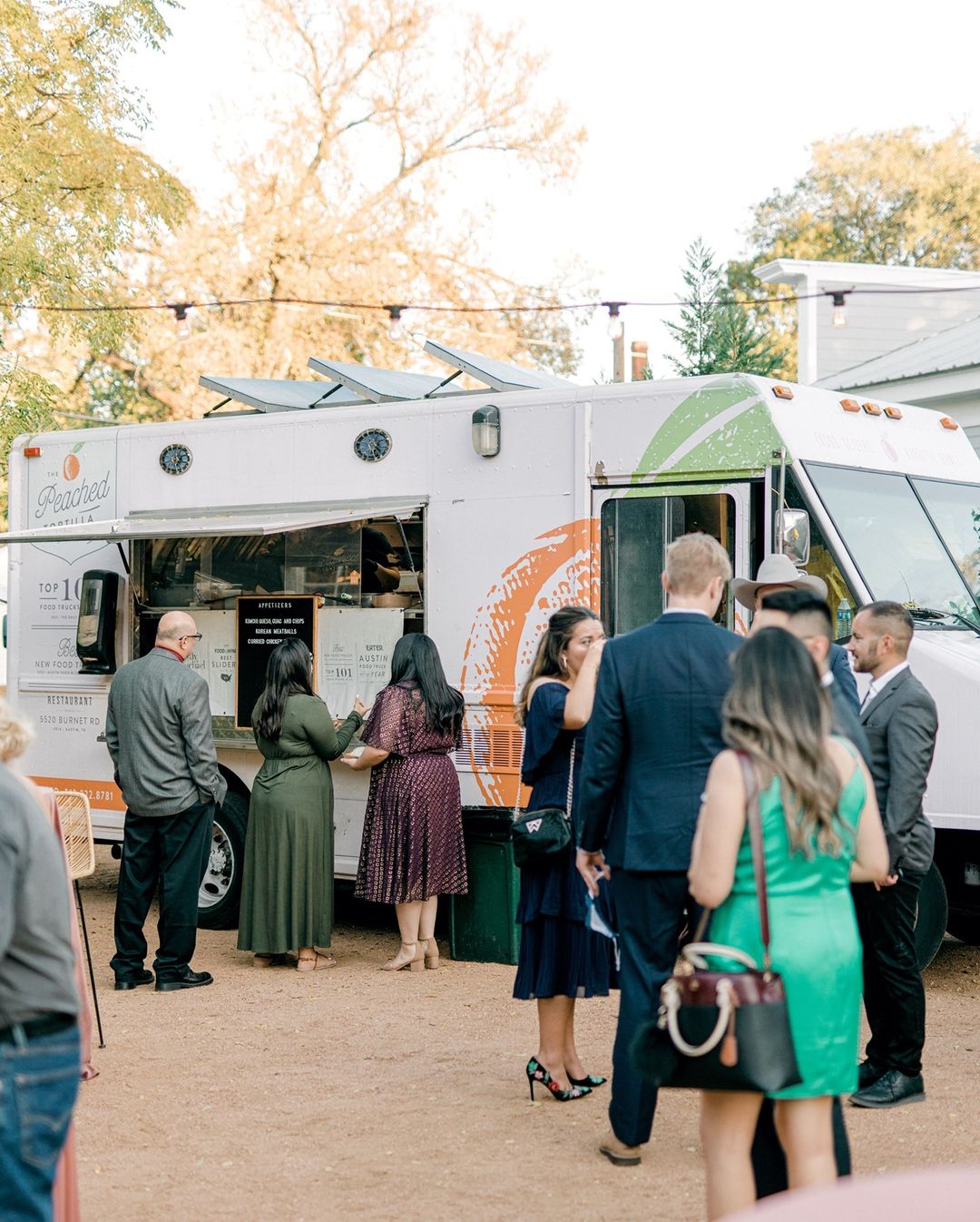 A group of well-dressed people stand in line at a food truck with the logo "Peached" on its side. Tables and string lights decorate the outdoor setting, hinting at a casual gathering or event. Trees are visible in the background.