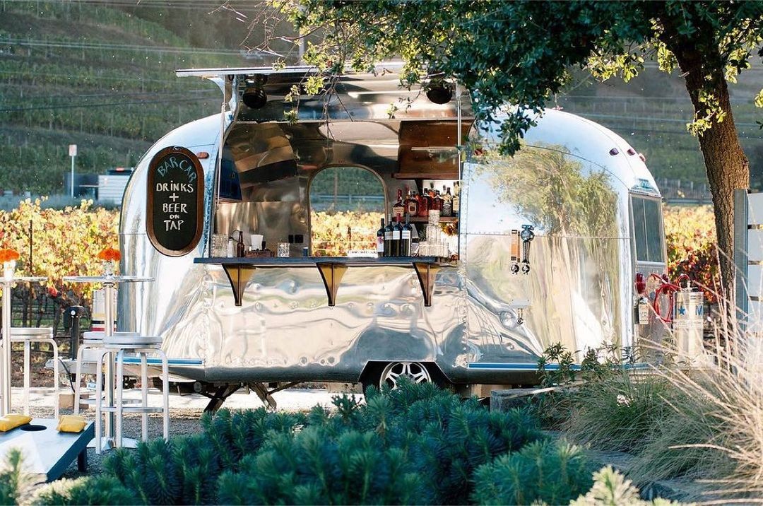 A shiny Airstream trailer converted into a mobile bar is parked outdoors near a vineyard. The bar window is open, displaying a variety of beverages on shelves and a chalkboard sign that reads "Drinks on Tap." Surrounding the area are plants and seating.