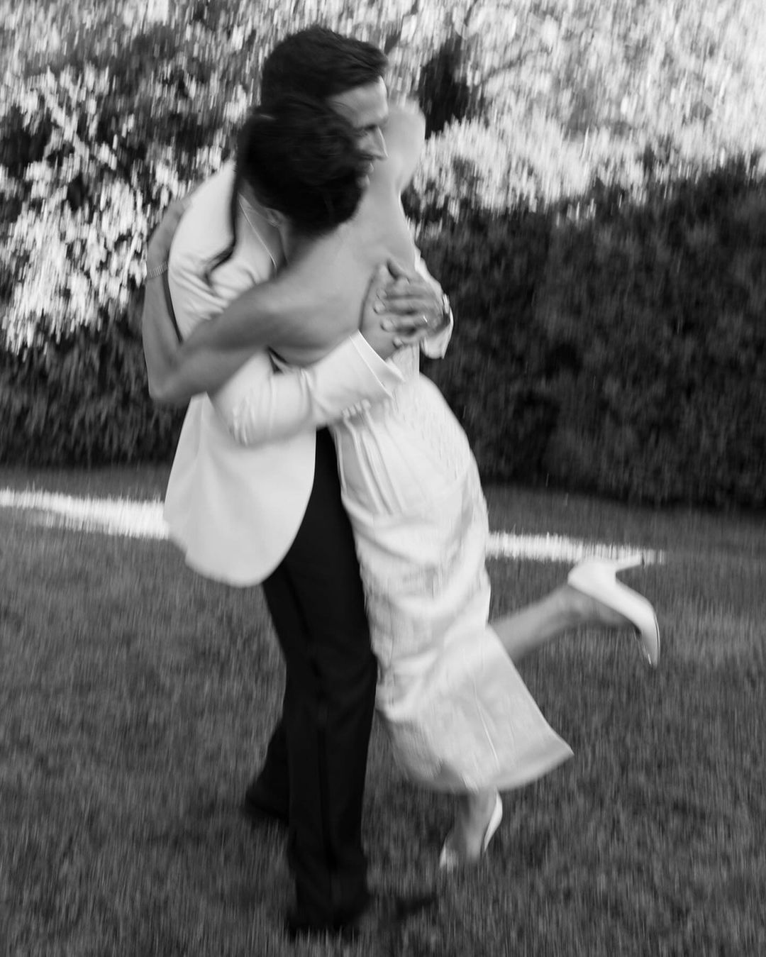 A couple dressed in formal wedding attire embraces tightly on a grassy area. The woman, in a strapless wedding dress and heels, is lifted off the ground by the man in a suit. The background is blurred, emphasizing the couple's joyous moment.