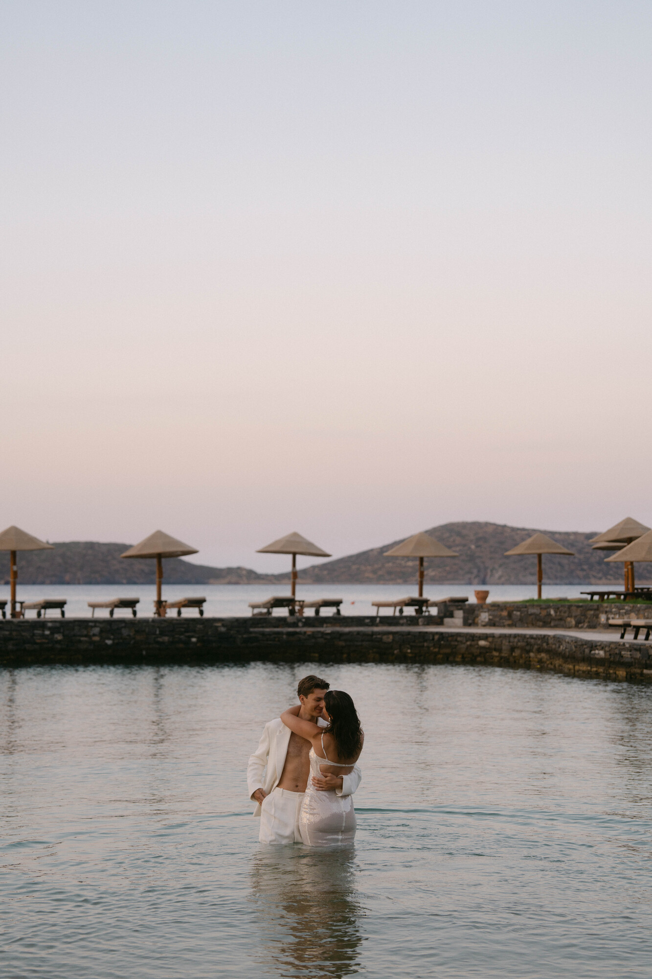 A couple stands in shallow water with the man wearing white trousers and an open shirt, and the woman in a white dress. They embrace and face each other affectionately. In the background, there are beach umbrellas, loungers, and a serene sky over distant hills.