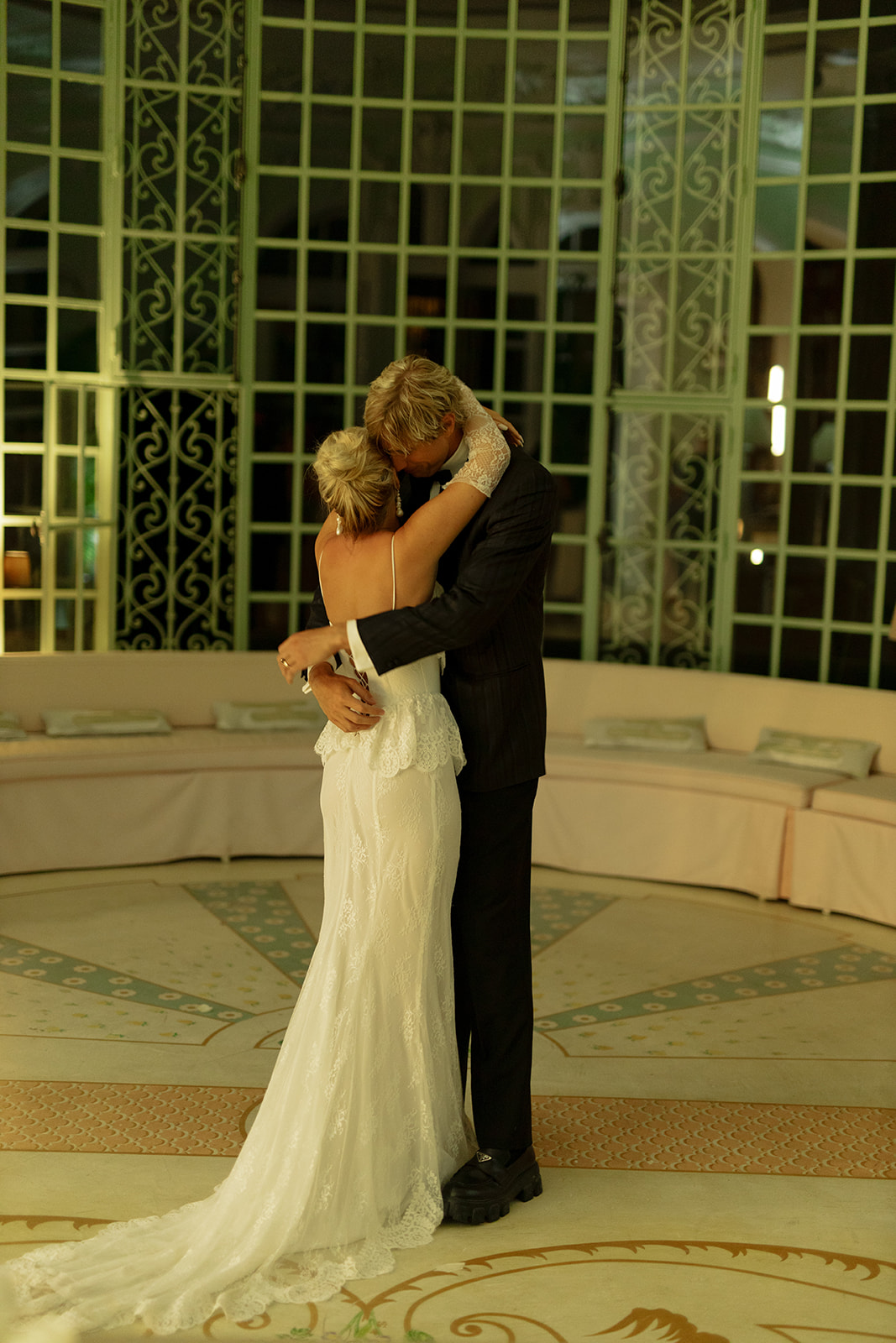 A couple shares a romantic kiss while hugging in an elegant, opulent room. The woman, wearing a white lace wedding dress, and the man, in a dark suit, are the focal points of the scene. They stand on a patterned floor with an ornate backdrop of decorative window panes.