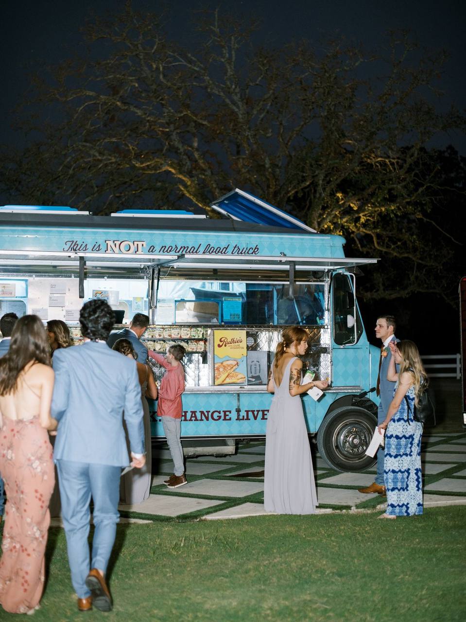 People are gathered in front of a blue food truck at night, chatting and waiting for their orders. The truck has lights illuminating its counter and a sign that partially reads, "This is NOT a normal food truck." Trees and dim lighting set the ambiance.